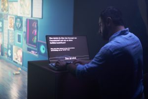 Eine Person steht in einem dunklen Raum vor einem Computer. Sie tippt etwas ein. Auf dem Bildschirm wird eine Frage über Freundschaft gestellt.